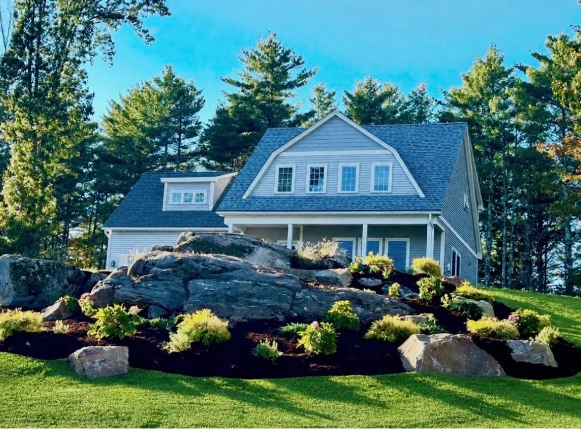 Backyard design with large natural boulder retaining wall and layered plantings on a sloped front yard - Up North Scapes NH
