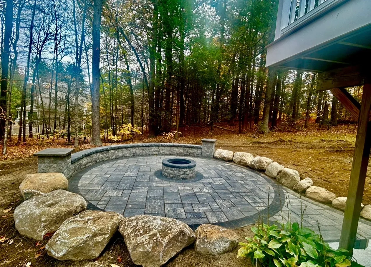 Backyard design with curved stone-clad seat wall, built-in fire pit, and boulder-lined patio - Up North Scapes NH