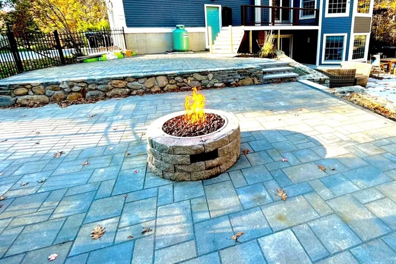 Paver patio with a fire pit below a stone faced wall behind a blue house, built by Up North Scapes