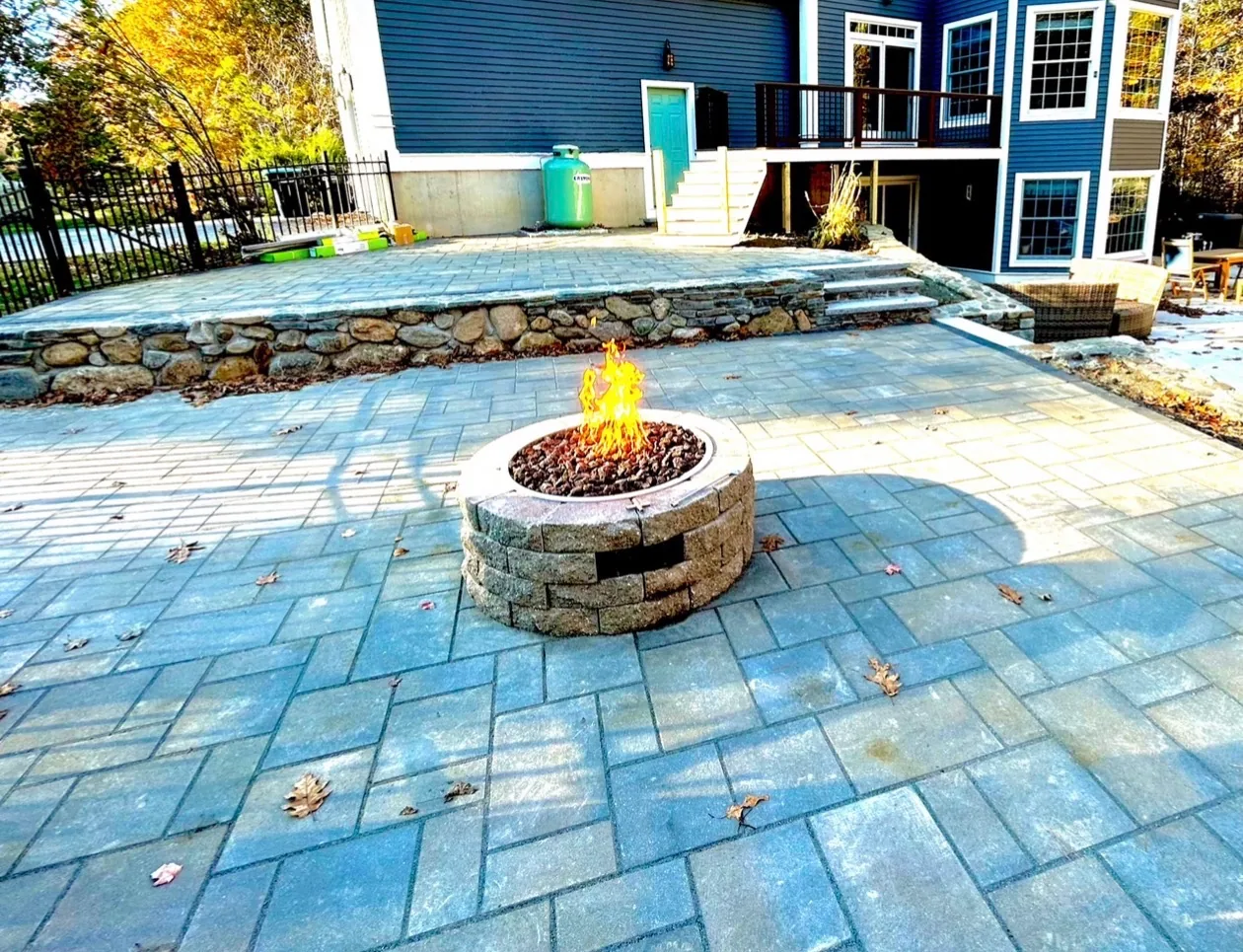 Backyard design with elevated paver patio, stone-faced retaining wall, and built-in fire pit at dusk - Up North Scapes NH