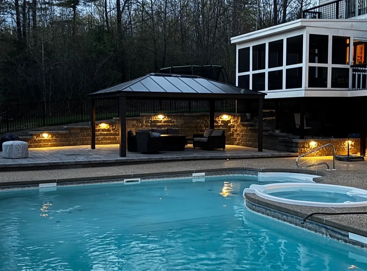Backyard design with pool, hot tub, and lighted pergola-covered lounge area at dusk - Up North Scapes NH