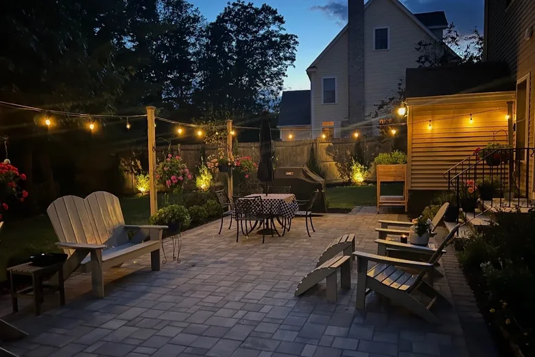 Paver patio at dusk under string lights, with lit plantings along the house, built by Up North Scapes