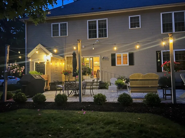 Paver patio behind a house at twilight with string lights on posts