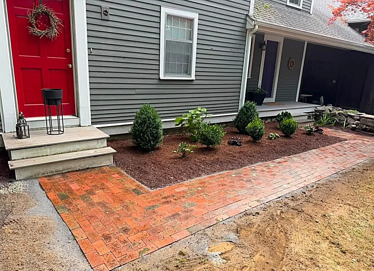 Red brick walk turning the corner in front of a grey colonial with a red door