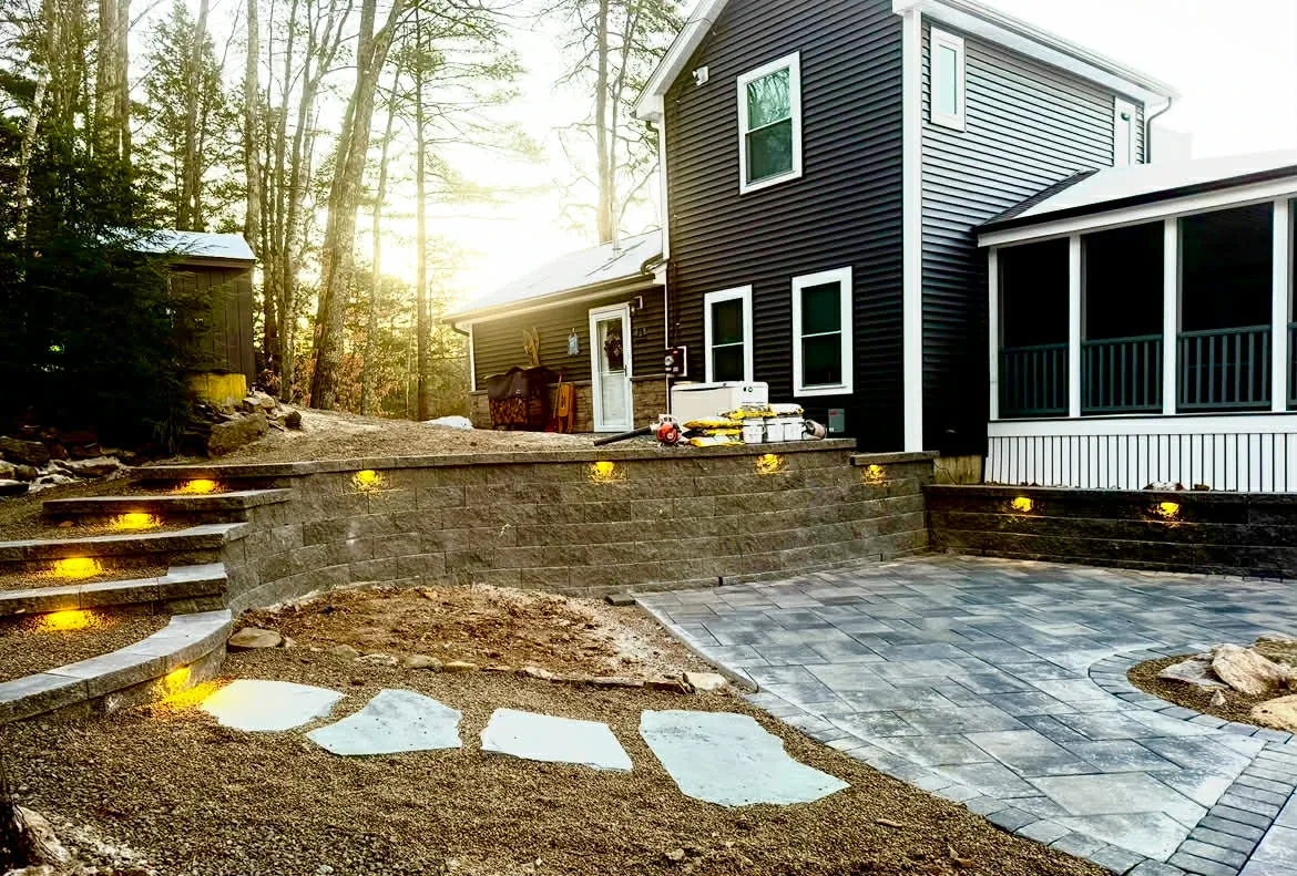 Block retaining wall with integrated step and face lighting beside a paver patio at dusk, New Hampshire Seacoast