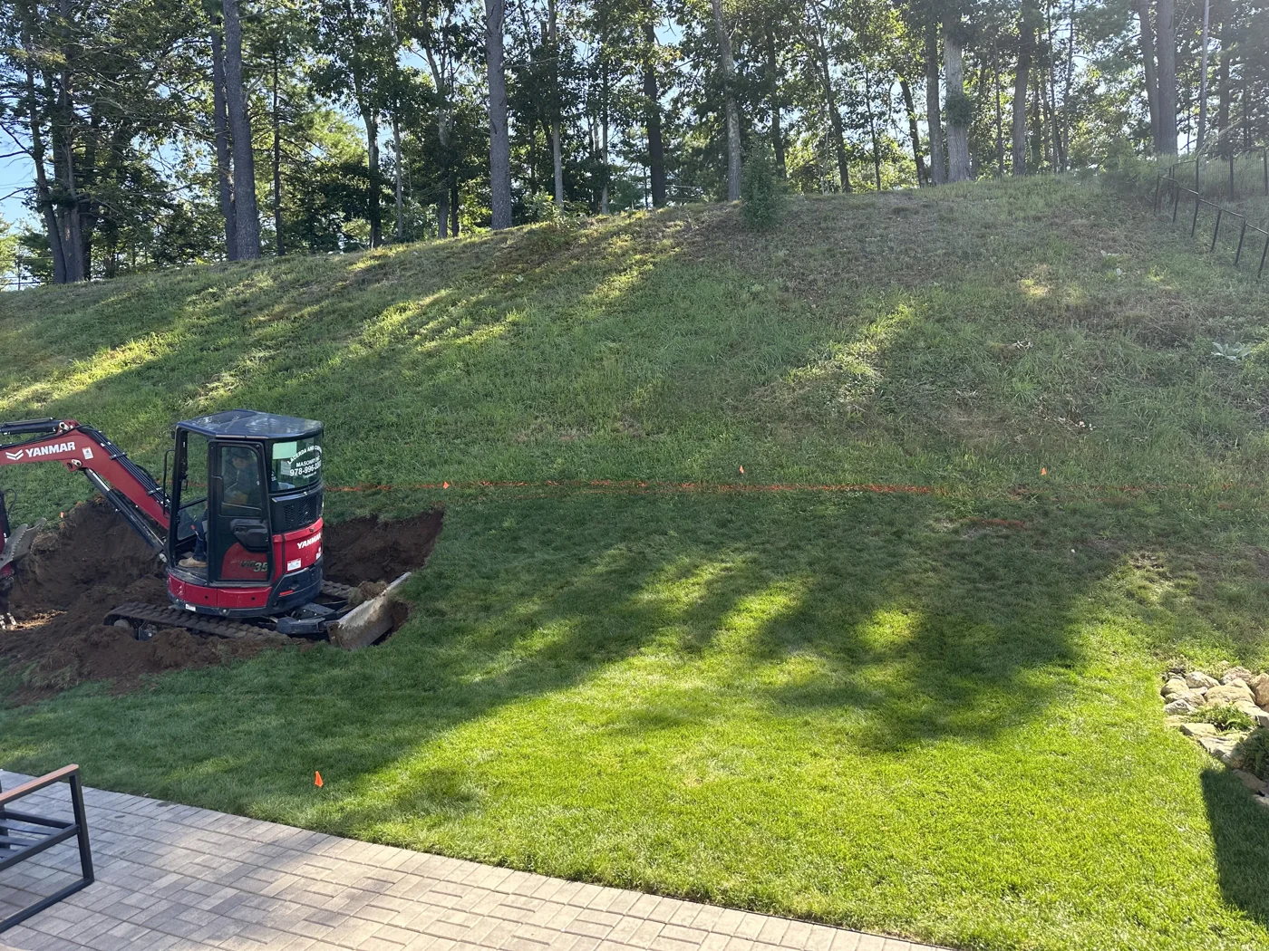 Excavator starting to cut into a steep grassy hill behind a backyard patio