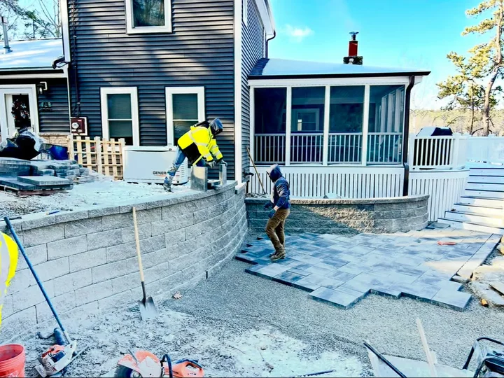Two workers in winter jackets setting pavers beside a new block wall behind a house