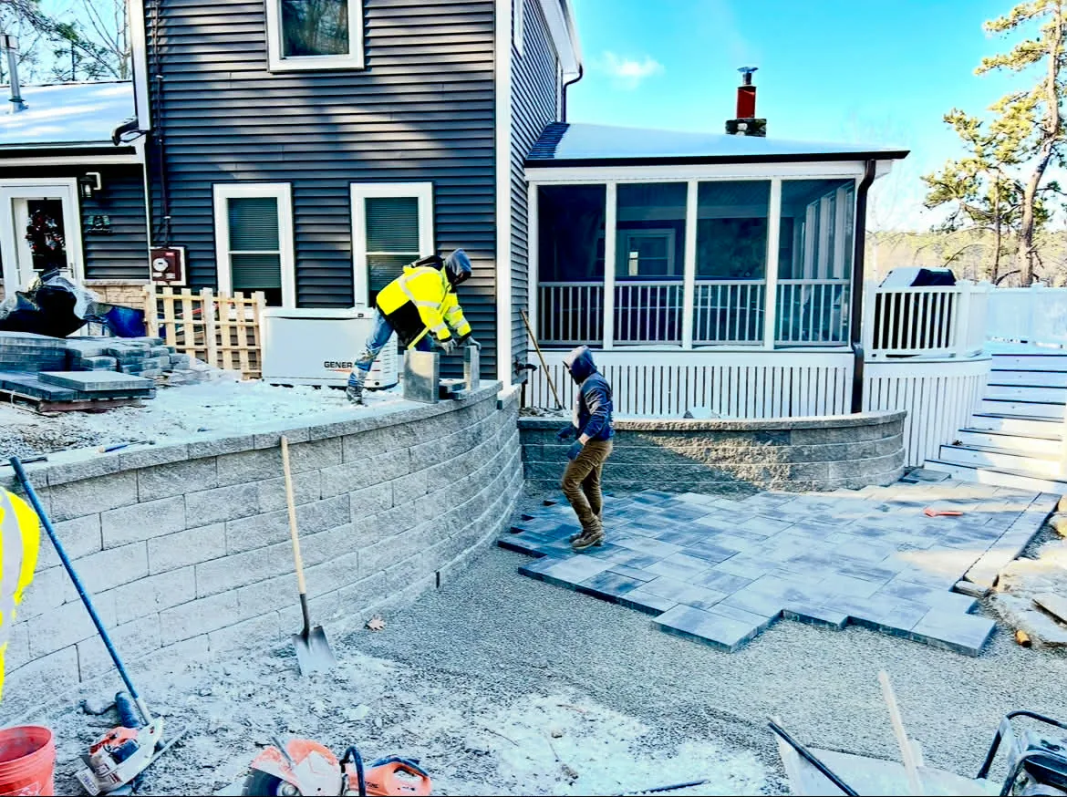 Two workers in winter jackets setting pavers beside a new block wall behind a house